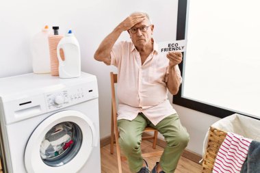 Üst düzey bir adam elinde çevre dostu kağıtlarla çamaşır yıkıyor. Kafası karışık, şaşırmış ve sinirli bir yüz ifadesi var. 