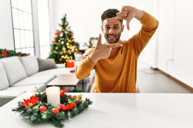 Arap genç adam Noel ağacının yanında oturmuş gülümsüyor ve elleri ve parmakları gülen yüzlü bir çerçeve yapıyor. yaratıcılık ve fotoğrafçılık kavramı. 