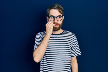 Çizgili tişört ve gözlük takan beyaz sakallı adam gergin ve elleri ağızda tırnak yerken gergin görünüyor. anksiyete. 