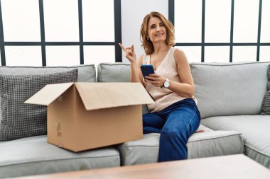 Orta yaşlı bir kadın kanepede oturmuş telefonda teslimat paketini kontrol ediyor. Gülümsüyor, mutlu bir şekilde elini ve parmağını yana doğru gösteriyor. 