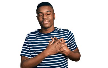 Sıradan çizgili tişört giyen, elleri göğsünde gülümseyen, gözleri kapalı, yüzünde minnet ifadesi olan genç bir Afro-Amerikan. sağlık kavramı. 