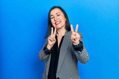 Orta yaşlı İspanyol kadın, iş kıyafetleri içinde kameraya bakıp zafer işareti yaparken gülümsüyor. İki numara.. 