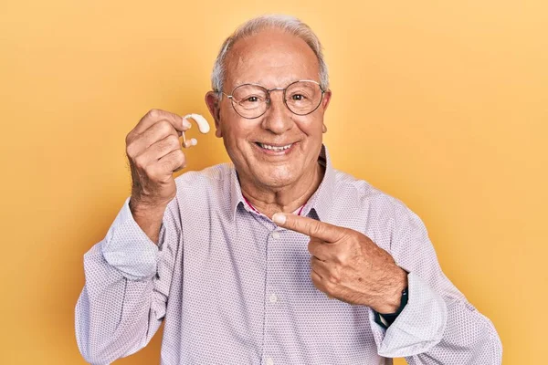 Senior man with grey hair holding medical hearing aid smiling happy pointing with hand and finger