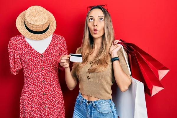 Beautiful hispanic woman holding shopping bags and credit card making fish face with mouth and squinting eyes, crazy and comical.