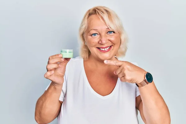 Middle age blonde woman holding cosmetic moisturizer facial cream smiling happy pointing with hand and finger