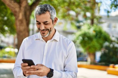 Orta yaşlı, kır saçlı, mutlu gülümseyen bir adam şehirde akıllı telefon kullanıyor..