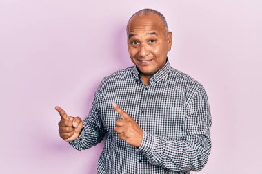 Orta yaşlı, sıradan kıyafetler giyen, gülümseyen ve kameraya bakan, iki eli ve parmakları yanda olan Latin bir adam.. 