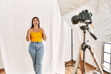 Fotoğrafçılık stüdyosunda model olarak poz veren genç, güzel İspanyol kadın şaşırmış ve şaşırmış bir şekilde yukarı bakıp parmaklarını göstermiş ve kollarını kaldırmıştı.. 