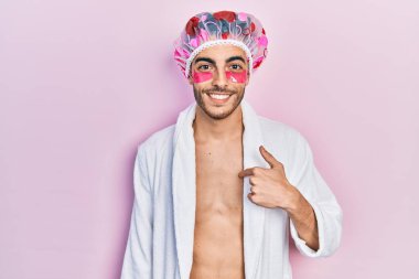 Bornoz ve duş bonesi takan genç İspanyol adam. Göz torbaları kullanıyor. Göz bantları takıyor, gülümseyen ve gururlu birini işaret ediyor. 