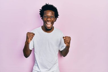 Beyaz bir tişört giyen genç Afro-Amerikalı adam kollarını kaldırıp, gözleri açık bir şekilde başarıyı kutluyor ve şaşırıyor. Kazanan konsept. 