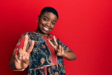 Afro-Amerikan tarzı giyinmiş genç bir kadın kameraya bakıp zafer işareti yaparken gülümsüyor. İki numara.. 