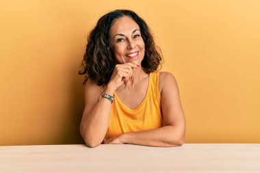 Orta yaşlı, sıradan kıyafetler giyen güzel bir kadın masaya oturmuş kameraya kendinden emin bir şekilde bakıyor kollarını kavuşturmuş ve çenesini kaldırmış gülümsüyor. Olumlu düşünmek. 