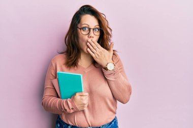 Kafkasyalı genç bir kadın elinde kitapla ağzını kapatıyor, şok geçiriyor ve hata yapmaktan korkuyor. şaşırmış ifade 