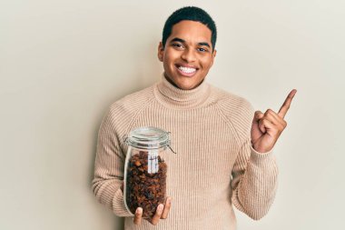 Genç, yakışıklı, elinde kuru üzümlerle dolu bir kavanoz tutan ve mutlu bir şekilde elini ve parmağını yana doğru gösteren bir adam. 