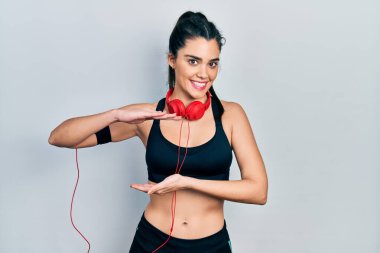 Spor kıyafetleri giyen, kulaklık takan, büyük ve büyük beden işareti gösteren İspanyol bir kız. Kameraya bakıp gülümsüyor. ölçüm kavramı. 