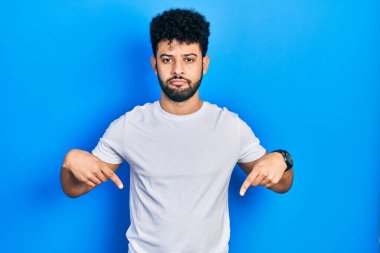 Sakallı, beyaz tişörtlü genç Arap adam üzgün ve üzgün görünüyor, parmaklarıyla yönü gösteriyor, mutsuz ve depresif.. 
