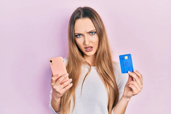 Młoda Blondynka Trzyma Smartfon Kartę Kredytową Szoku Twarzy Patrząc Sceptyczny — Zdjęcie stockowe