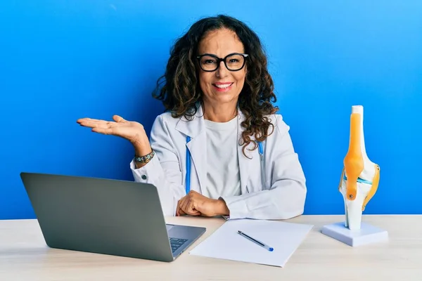 Beautiful middle age woman doctor at orthopedic clinic smiling cheerful presenting and pointing with palm of hand looking at the camera.