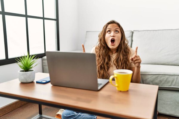 Красивая латиноамериканка с помощью ноутбука дома удивлена и удивлена, глядя вверх и указывая пальцами и поднятыми руками. 