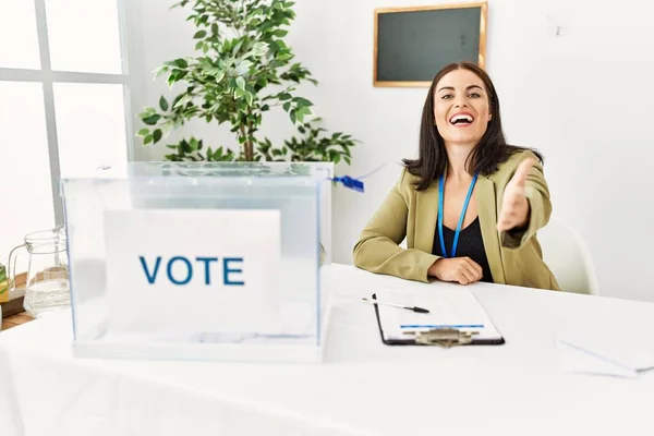 投票投票で選挙テーブルに座っている若いブルネットの女性は挨拶と歓迎として握手を提供する優しい笑顔 成功したビジネス — ストック写真