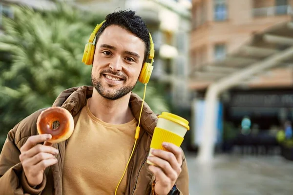 若いヒスパニック系の男が街でヘッドフォンを使用して朝食を持っています — ストック写真