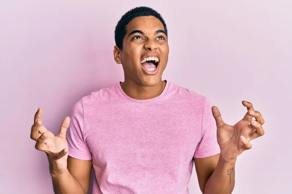 カジュアルなピンクのTシャツを着た若いハンサムなヒスパニック系の男が狂気と怒鳴り 積極的な表現と腕を上げて叫びました フラストレーションコンセプト — ストック写真