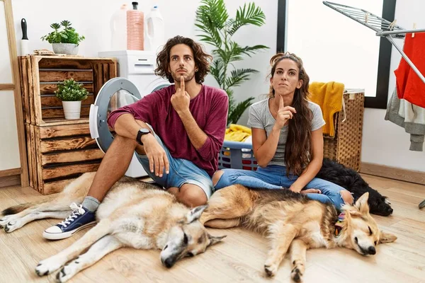 Young hispanic couple doing laundry with dogs thinking concentrated about doubt with finger on chin and looking up wondering