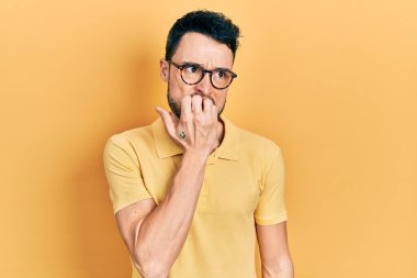Günlük kıyafetler ve gözlük takan genç İspanyol adam gergin ve gergin görünüyor. Elleri ağızda, tırnaklarını yiyor. anksiyete. 