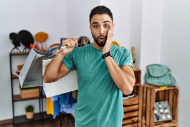 Satış mağazasında sakallı, alışveriş torbaları tutan genç yakışıklı adam korkmuş ve şok olmuş, şaşırmış ve şaşırmış bir halde. 