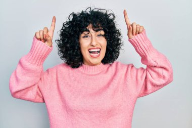 Orta Doğulu genç bir kadın gündelik kıyafetler giyiyor. Şaşırmış, şaşırmış, parmaklarıyla işaret ediyor ve kollarını kaldırıyordu.. 