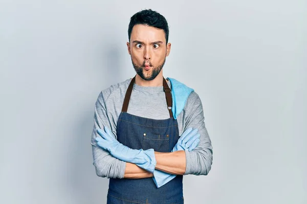 Young hispanic man wearing apron with arms crossed making fish face with mouth and squinting eyes, crazy and comical.