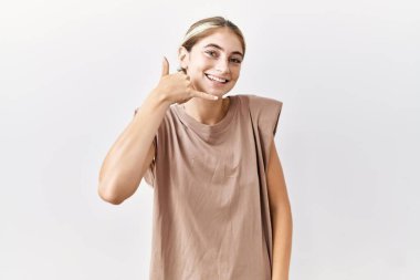 İzole edilmiş arka planda duran genç sarışın kadın gülümseyerek el ve parmaklarıyla telefon hareketi yapıyor telefonda konuşuyor gibi. iletişim kavramları. 