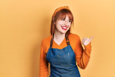 Önlük takan, mutlu yüzlü, gülümseyen ve baş parmağıyla yan tarafı gösteren kızıl saçlı genç bir kadın.. 