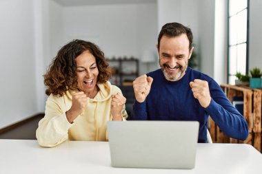 Orta yaşlı, yumrukları havada mutlu gülümseyen bir İspanyol çift. Evde dizüstü bilgisayarla masada oturmak..
