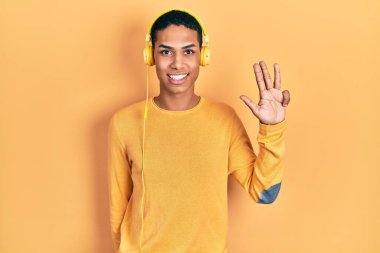 Genç Afro-Amerikalı adam müzik dinlerken kulaklık takıyor ve dört numaralı parmaklarıyla işaret ederken kendinden emin ve mutlu gülümsüyor.. 