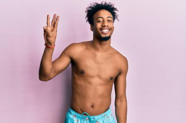 Sakallı, mayo giymiş, üç numaralı parmaklarıyla gösteriş yapan, kendinden emin ve mutlu genç Afrikalı Amerikalı bir adam.. 