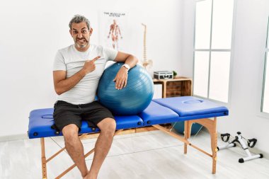 Orta yaşlı İspanyol adam ağrı kesici kliniğinde elinde pilates topu tutarken endişeli ve endişeli bir ifade sergiliyor. 