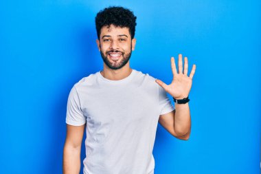 Sakallı, beyaz tişörtlü genç bir Arap. Beş numaralı parmaklarıyla kendini beğenmiş, mutlu bir şekilde gülümsüyor.. 