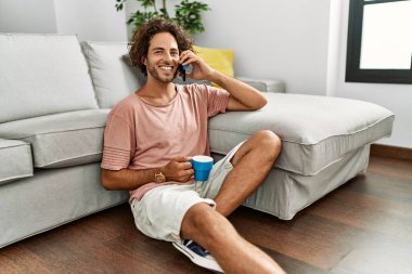 Genç İspanyol adam kahve içiyor ve akıllı telefonla konuşuyor. Evde yerde oturuyor..