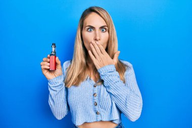 Genç beyaz kadın elinde elektronik sigarayla ağzını eliyle kapatıyor, şokta ve hata yapmaktan korkuyor. şaşırmış ifade 