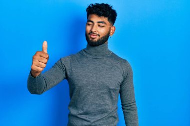 Balıkçı yaka kazak giyen sakallı bir Arap gururlu görünüyor, gülümsüyor ve baş parmak hareketi yapıyor. 