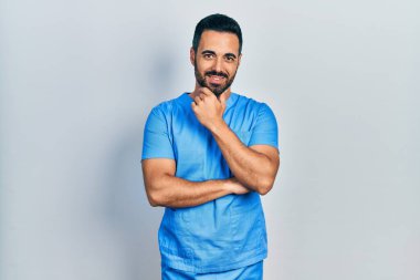 Sakallı yakışıklı, mavi erkek hemşire üniforması giyen kolları çapraz ve çenesi açık kameraya gülümseyen bir İspanyol. Olumlu düşünmek. 