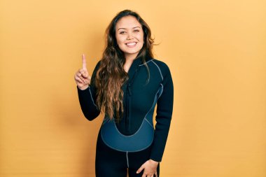 Genç İspanyol kız dalgıç neoprene üniforması giyiyor ve bir numaralı parmağını gösterip işaret ederken kendinden emin ve mutlu gülümsüyor.. 