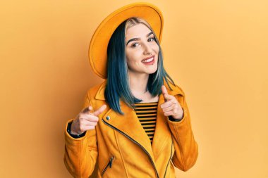 Sarı şapkalı ve deri ceketli genç modern kız mutlu ve komik yüzlü parmaklarını kameraya doğrultuyor. iyi enerji ve titreşimler. 