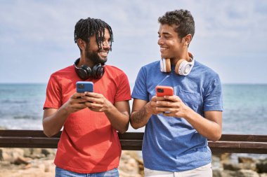 Sahilde akıllı telefon ve kulaklık kullanarak gülümseyen iki Afrikalı Amerikalı adam..