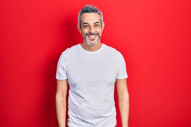 Yakışıklı, orta yaşlı, gri saçlı, sıradan beyaz tişörtlü, pozitif ve mutlu görünen, kendine güvenen, gülümseyen, dişlerini gösteren bir adam. 