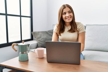 Genç esmer kadın evde dizüstü bilgisayar kullanıyor. Bir fincan kahve içiyor. Yüzünde gülümsemeyle yan yana bakıyor. Kendinden emin gülen. 