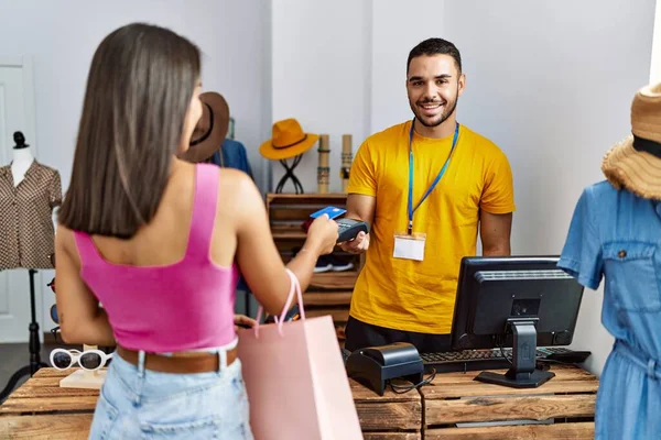 Young latin customer man paying to shopkeeper using credit card at clothing store.