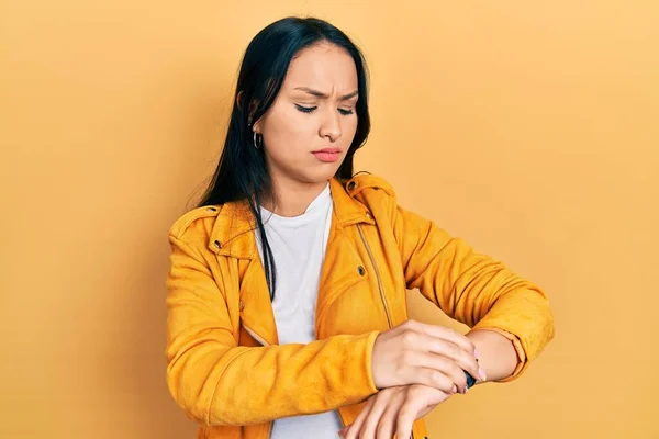 Beautiful hispanic woman with nose piercing wearing yellow leather jacket checking the time on wrist watch, relaxed and confident