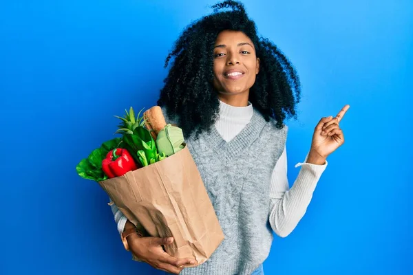 アフリカ系アメリカ人女性とアフロヘア保持紙袋で食料品笑顔幸せなポインティングで手と指で側面 — ストック写真